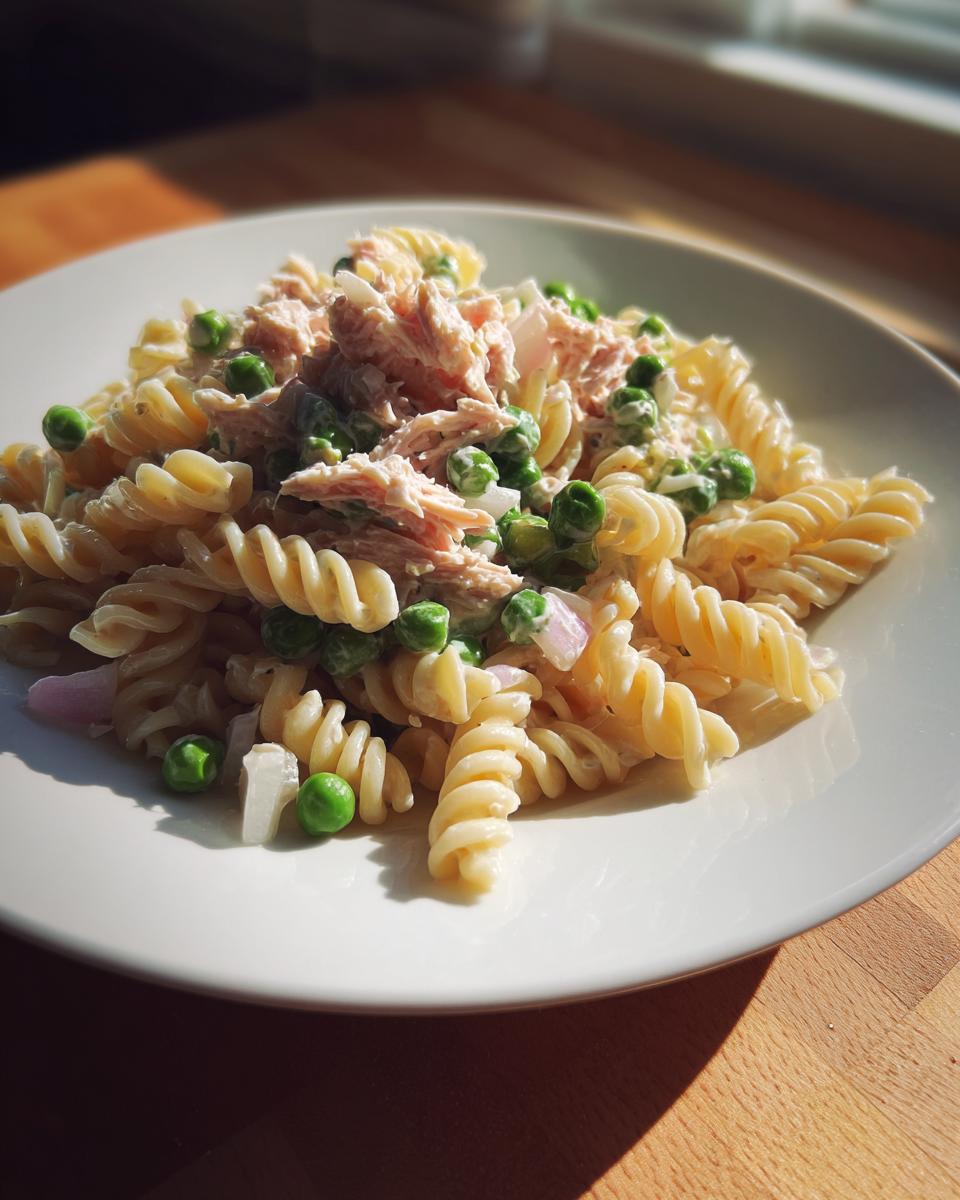 A close-up of a serving of creamy tuna pasta salad made with rotini pasta, green peas, and red onion.
