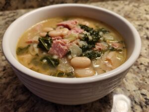 Close-up of a white bowl filled with creamy white bean soup featuring chunks of ham, kale, and white beans.