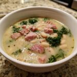 A close-up of a hearty white bean soup with chunks of ham, kale, and grated cheese in a white bowl.