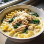 A close-up of a white bowl filled with creamy white chicken lasagna soup, topped with shredded chicken, spinach, and Parmesan cheese.