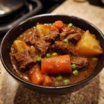 Close-up of a hearty bowl of crockpot beef stew featuring tender beef chunks, carrots, potatoes, and peas in a rich brown gravy.