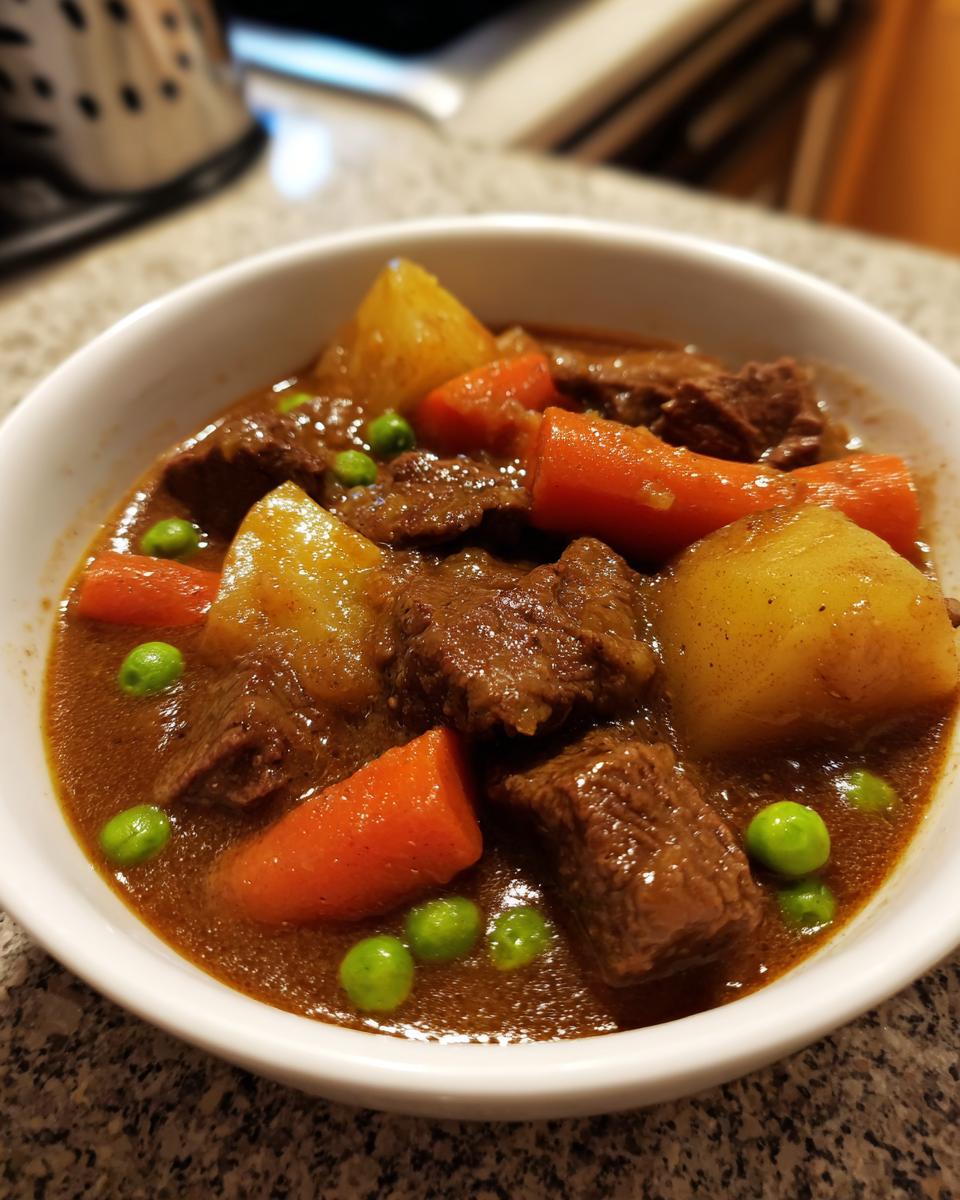 A close-up of a white bowl filled with rich crockpot beef stew featuring tender beef chunks, potatoes, carrots, and bright green peas.