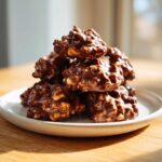 A tempting stack of homemade crockpot peanut clusters coated in glossy, melted chocolate, resting on a white plate.