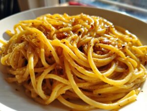 A close-up, bright photo of a serving of glossy, golden garlic noodles tossed with spices and garlic bits on a white plate.