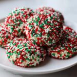 A stack of delicious christmas sprinkle cookies completely covered in red, green, and white nonpareils on a white plate.