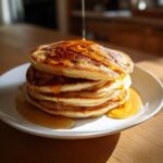 A tall stack of fluffy banana pancakes being drizzled with golden syrup on a white plate.