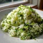 A close-up mound of fluffy cilantro lime rice, bright green with fresh herbs, served on a white plate.