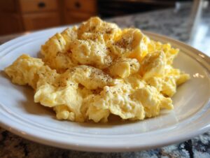 A close-up of fluffy, creamy scrambled eggs seasoned with cracked black pepper on a white plate.