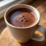 Close-up of rich, dark french hot chocolate with a layer of small bubbles on top, served in a beige mug.
