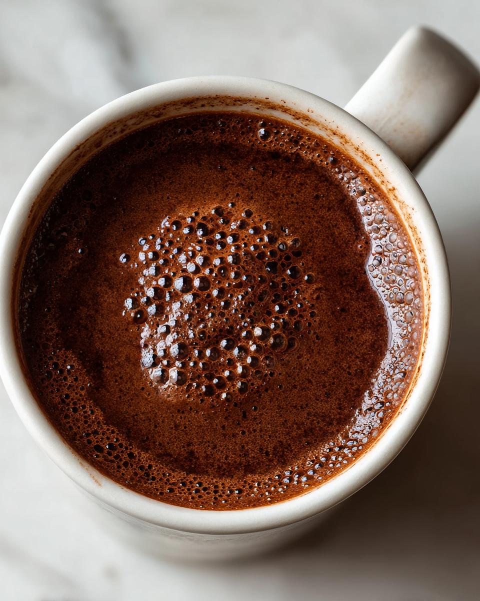 Overhead close-up of rich, dark french hot chocolate with a layer of small bubbles on top, served in a white mug.