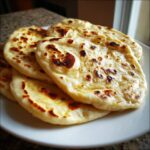 A close-up stack of fluffy, freshly made naan bread with golden-brown charred spots served on a white plate.