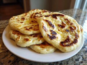 A stack of fluffy, freshly cooked naan bread brushed with butter, showing charred spots, served on a white plate.