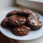 A pile of freshly baked, fudgy brownie cookies with cracked, shiny tops resting on a white plate.
