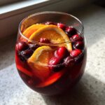 Close-up of a glass filled with vibrant red Thanksgiving punch, garnished with orange slices and whole cranberries.