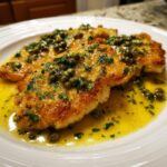 Close-up of two golden, pan-fried chicken piccata cutlets swimming in a bright lemon butter sauce topped with capers and parsley.
