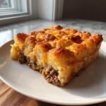 A square slice of baked tater tot casserole with a crispy, cheesy top layer resting on a white plate.
