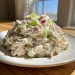 A close-up of a creamy mound of greek yogurt chicken salad mixed with diced celery and red onion on a white plate.