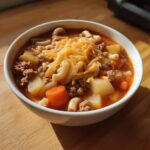 A close-up of a white bowl filled with rich hamburger soup, featuring ground beef, potatoes, carrots, macaroni, and topped with shredded cheddar cheese.