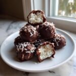 A stack of chocolate-covered almond joy truffles topped with shredded coconut, one is bitten open revealing the white coconut filling.