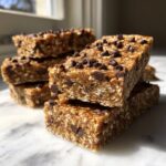 A stack of homemade, chewy protein bars studded with mini chocolate chips resting on a white marble surface.