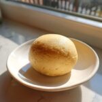 A smooth, light yellow ball of homemade marzipan recipe dough resting on a small white saucer near a window.