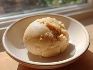 A scoop of light brown, smooth marzipan recipe dough resting in a small, light-colored ceramic bowl.