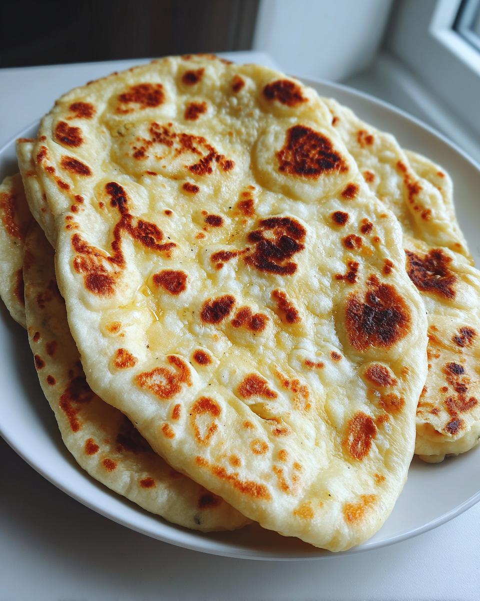 Close-up of several pieces of soft homemade naan bread stacked on a white plate, showing golden-brown charred spots.