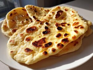 A stack of soft, freshly cooked homemade naan bread with golden brown charred spots on a white plate.