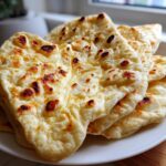 Close-up of a stack of freshly made, soft homemade naan bread with golden-brown bubbles and dark char marks.