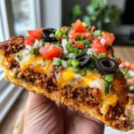 A hand holding a thick slice of Mexican pizza loaded with seasoned beef, melted cheese, black olives, tomatoes, and green onions.