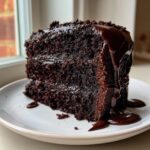 A rich, dark slice of moist chocolate cake recipe covered in dripping chocolate fudge sauce on a white plate.