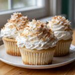 Three moist coconut cupcakes topped with white frosting and toasted coconut flakes sitting on a white plate.