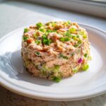A perfectly molded cylinder of creamy salmon salad topped with chopped green onions on a white plate.