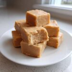 A stack of creamy, light brown peanut butter fudge squares piled on a white plate.