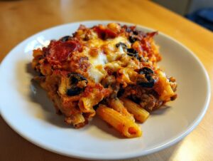 A close-up of a baked serving of pizza pasta featuring penne, melted cheese, pepperoni, and black olives.