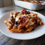 A serving of baked pizza pasta featuring penne, melted cheese, pepperoni slices, sausage, and black olives.