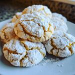 A close-up of several soft, yellow lemon crinkle cookies heavily dusted with powdered sugar on a light blue plate.