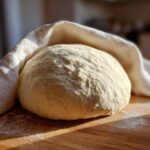 A perfectly proofed ball of pizza dough recipe resting on a floured wooden surface under a white cloth.