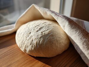 A perfectly risen ball of pizza dough recipe resting on a wooden surface, partially covered by a white kitchen towel.