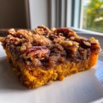 A close-up, sunlit slice of rich, orange pumpkin pecan cobbler topped with brown sugar crumble and whole pecans.