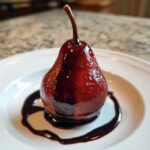 A single, deeply colored poached pear drizzled with dark syrup, served on a white plate.