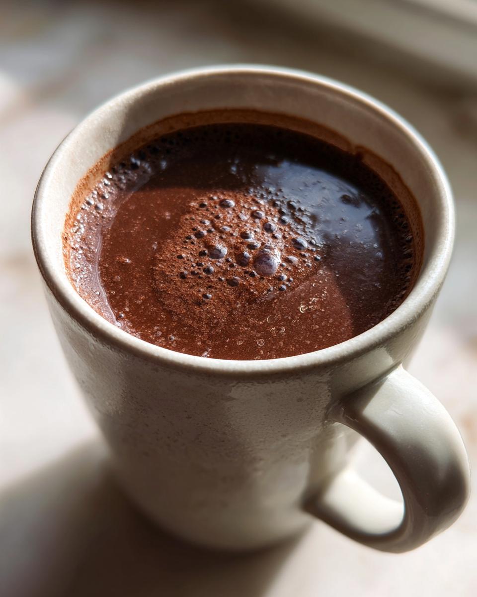 Close-up overhead shot of rich, dark french hot chocolate with small bubbles on the surface in a light ceramic mug.