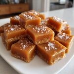 Close-up of several square, chewy, golden brown caramels sprinkled with flaky sea salt on a white dish.