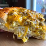 A close-up view of a thick slice of sausage gravy breakfast pizza showing melted cheese and eggs dripping.