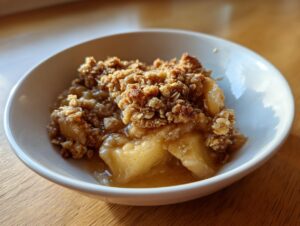 Close-up of a warm serving of apple crumble with gooey filling and crunchy oat topping in a white bowl.