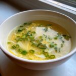 Close-up of a white bowl filled with hot egg drop soup, featuring wispy egg ribbons and chopped green onions.