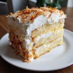 A tall slice of moist coconut poke cake layered with white frosting and topped with toasted coconut flakes.