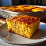 A golden-brown square slice of moist cornbread casserole served on a white plate.