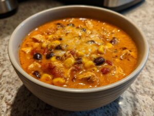 A close-up of a bowl filled with rich, orange slow cooker buffalo chicken chili, topped with melted cheese, corn, and black beans.