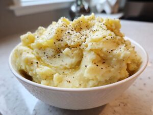 A white bowl overflowing with creamy slow cooker mashed potatoes, topped generously with cracked black pepper.